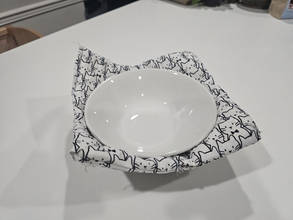 a cat fabric bowl cozy with a white bowl in it, on a white table